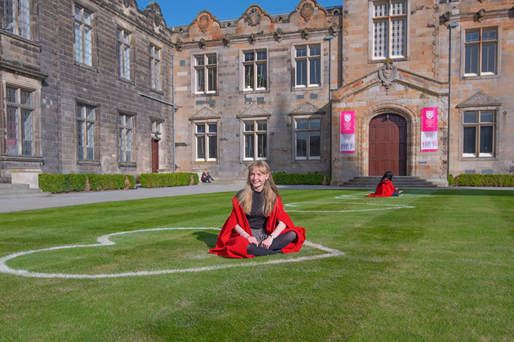A student sat on the grass in St Salvators quad, wearing a gown. A heart shape has been drawn on the grass, which the student is sitting inside.